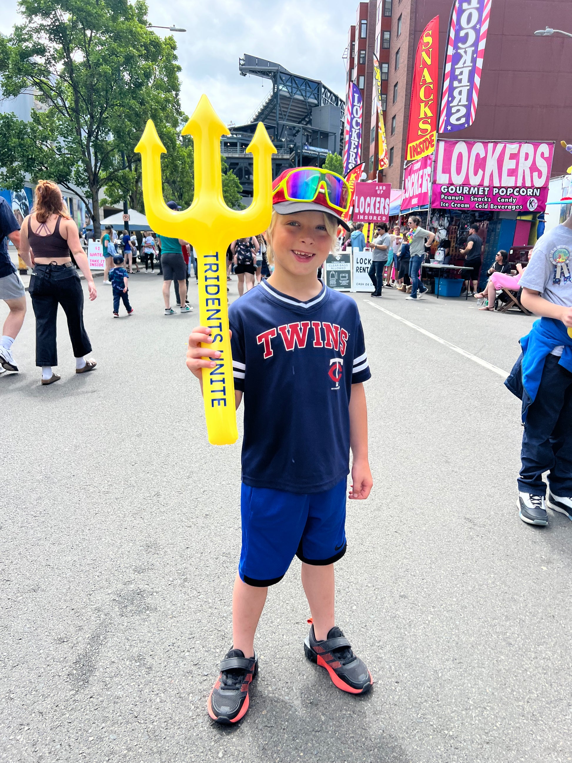 Child holding a yellow trident toy with colorful sunglasses, wearing a navy 'Twins' shirt and blue shorts, on a street with people and signs in the background. Seattle Mariners. Tridents Unite